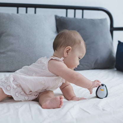 A baby reaching out to touch the rocking PlanToys wooden penguin wobbler on a soft bed.
