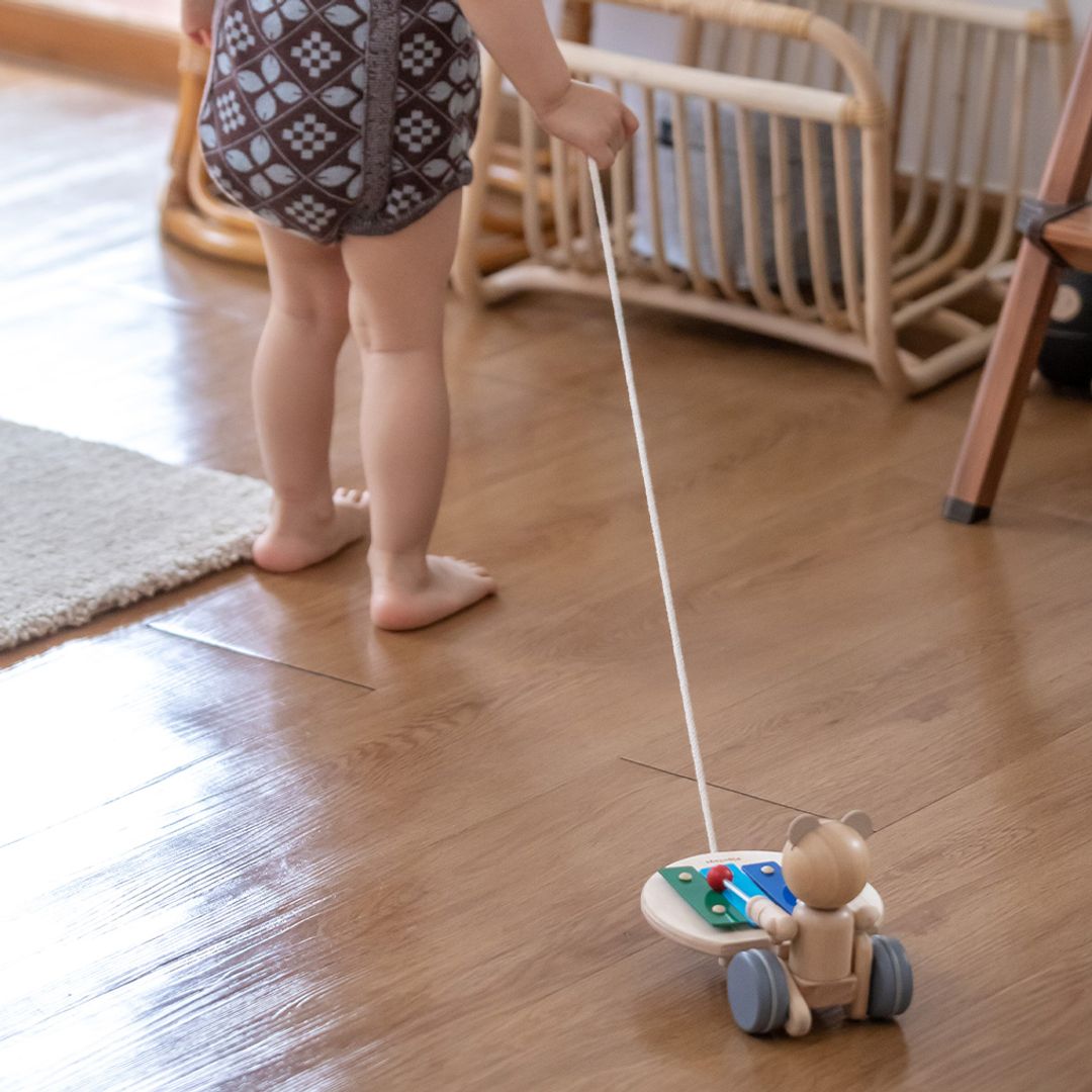 A toddler's legs walking across a wooden floor while towing the cheerful PlanToys pull along musical bear.