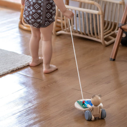 A toddler's legs walking across a wooden floor while towing the cheerful PlanToys pull along musical bear.