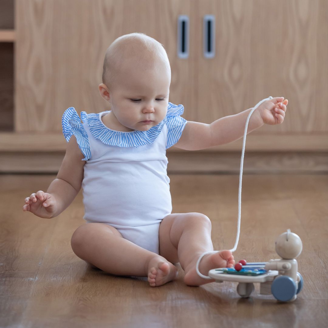 A baby in a blue-trimmed white onesie sitting on the floor and grasping the pull cord of the PlanToys musical bear.