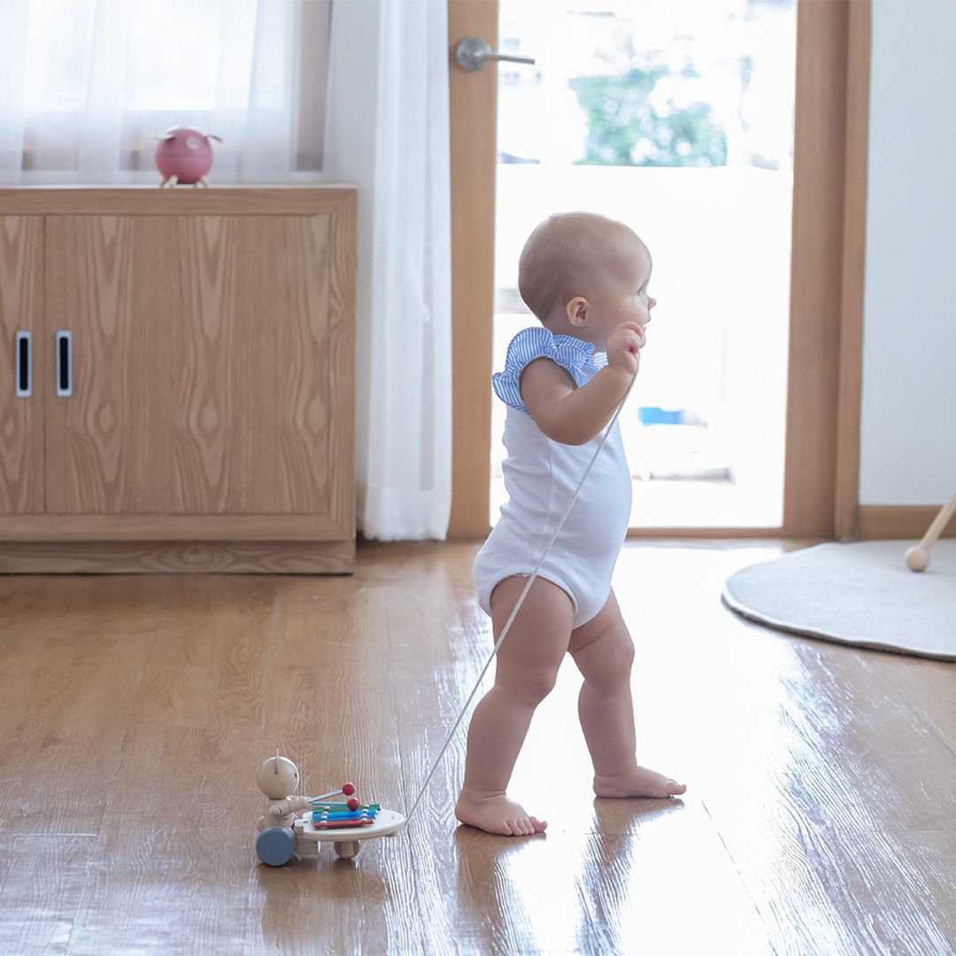 A baby practicing walking while pulling the earth-friendly PlanToys musical bear toy across a sunny room.