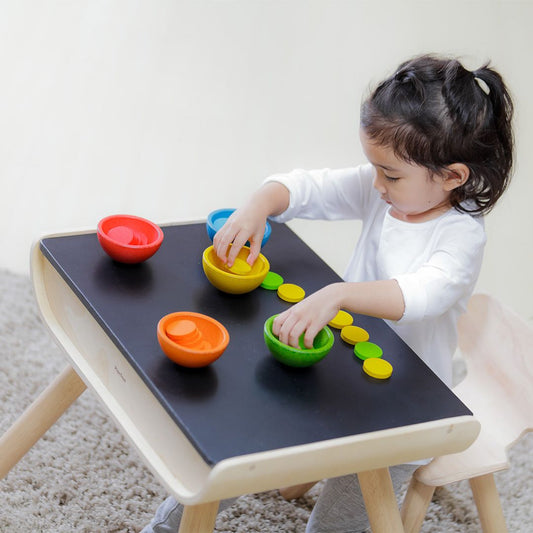 A toddler developing early math skills by sorting colorful wooden coins into the matching PlanToys cups.