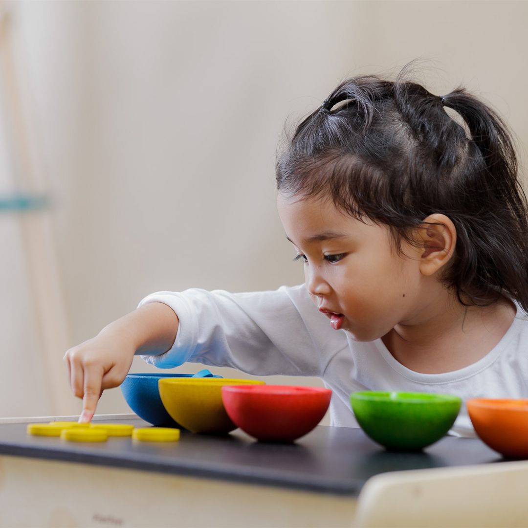 A young girl practicing her counting skills by pointing at the non-toxic yellow wooden coins from the PlanToys learning set.