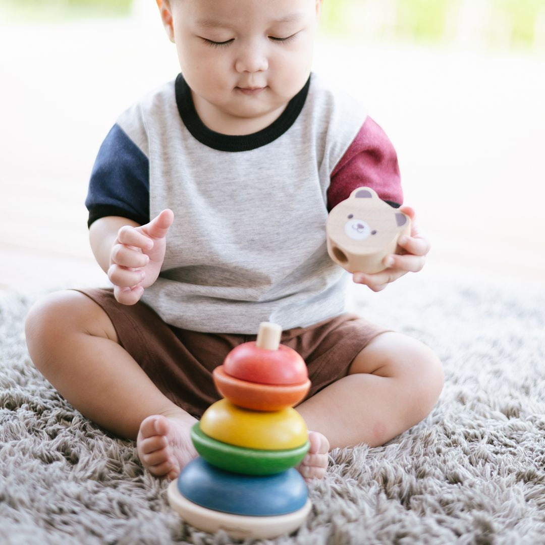A baby sitting on a soft rug playing with the fully assembled PlanToys wooden bear stacking ring toy.