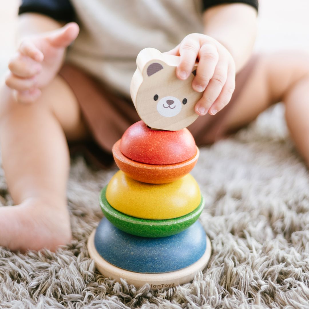 Close-up of a child's hand placing the wooden bear head on top of the colorful PlanToys stacking rings.