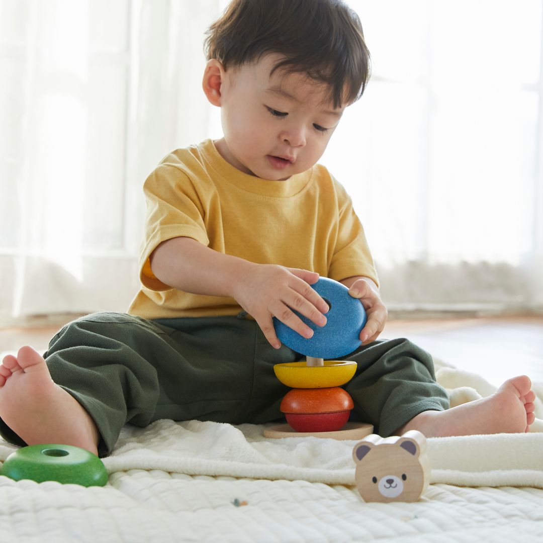 A toddler carefully sliding a blue wooden ring onto the rod of the PlanToys eco-friendly stacking toy.
