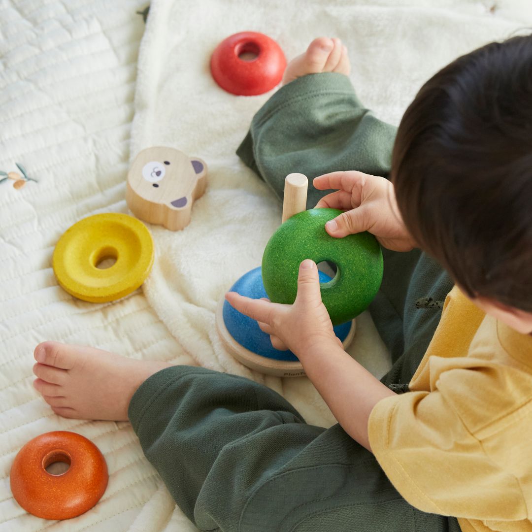 A toddler sitting on a blanket surrounded by colorful wooden rings, preparing to stack the green one.