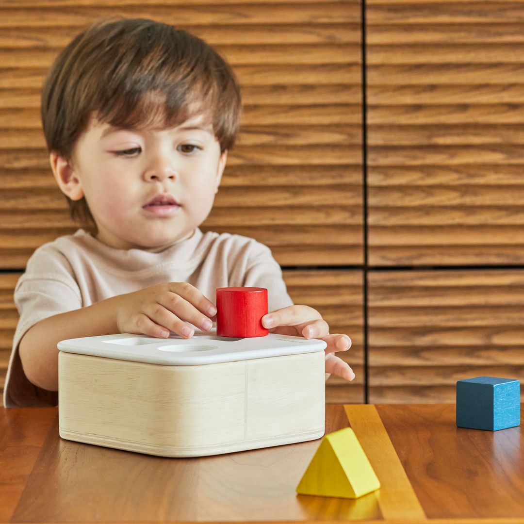 Close-up of a child's hand aligning a yellow star block with the corresponding opening on the non-toxic PlanToys wooden lid.