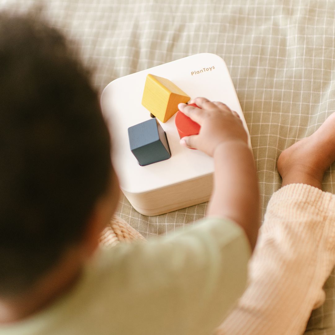 Detailed lifestyle shot of a toddler successfully matching the red square block to the PlanToys wooden shape sorter.