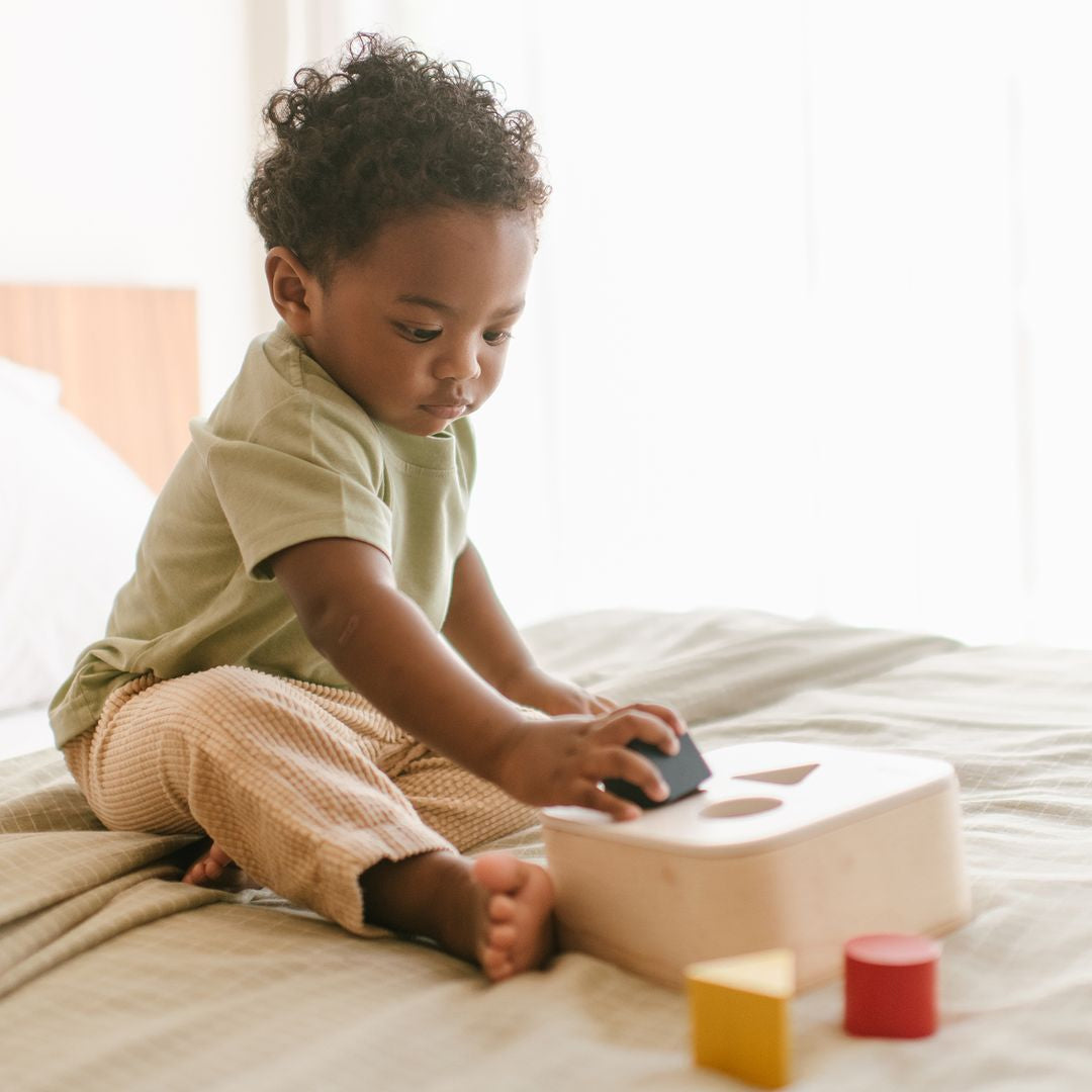 A happy toddler girl smiling while holding the green triangle and red square blocks from her PlanToys sorting set.