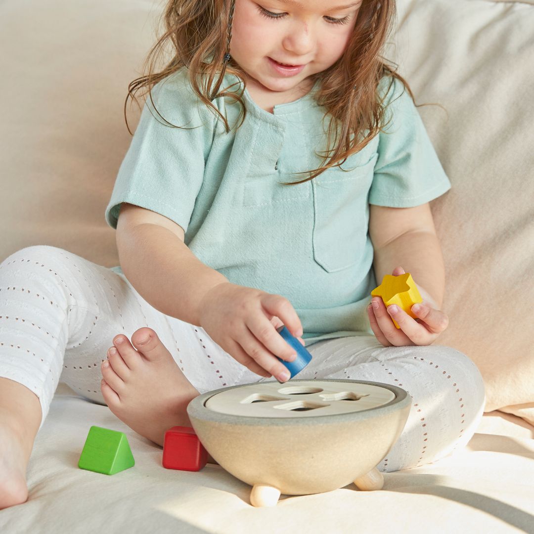 A young girl practicing fine motor skills by placing a blue circular block into the sustainable PlanToys shape sorter.