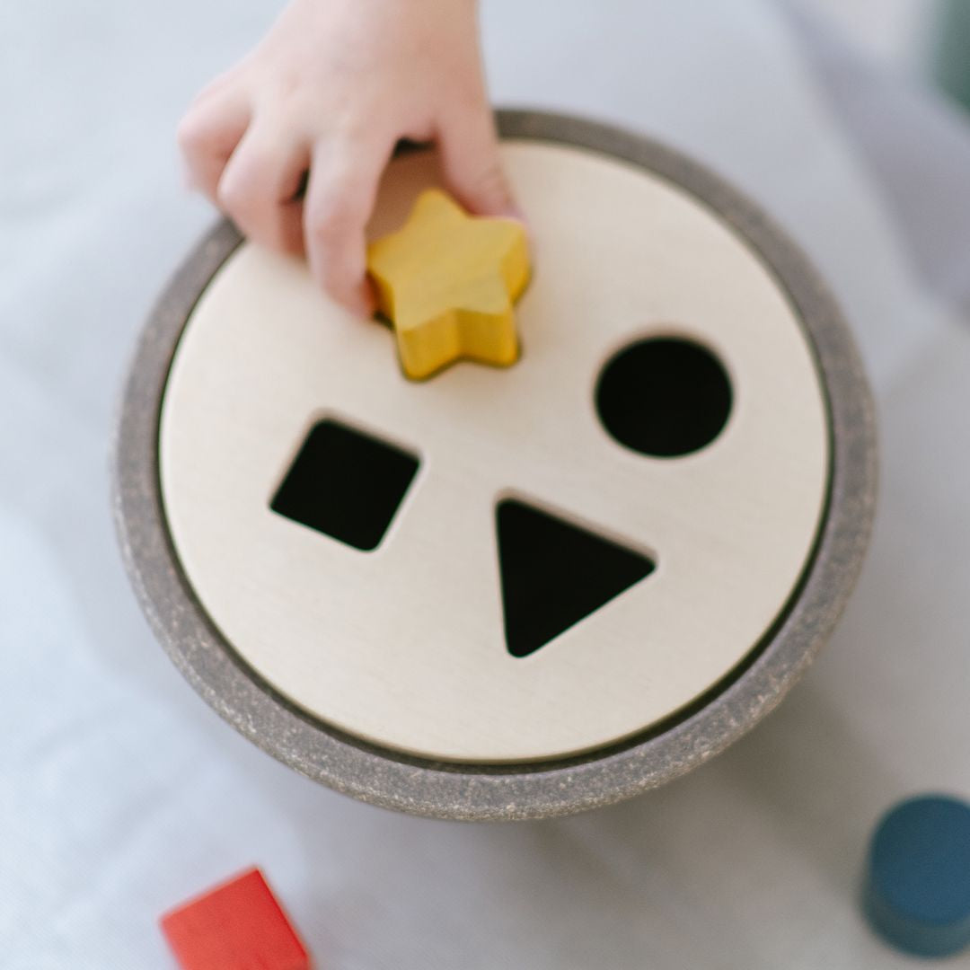 Close-up of a child's hand aligning a yellow star block with the corresponding opening on the non-toxic PlanToys wooden lid.