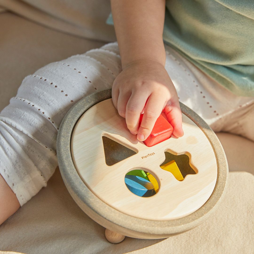 Detailed lifestyle shot of a toddler successfully matching the red square block to the PlanToys wooden shape sorter.