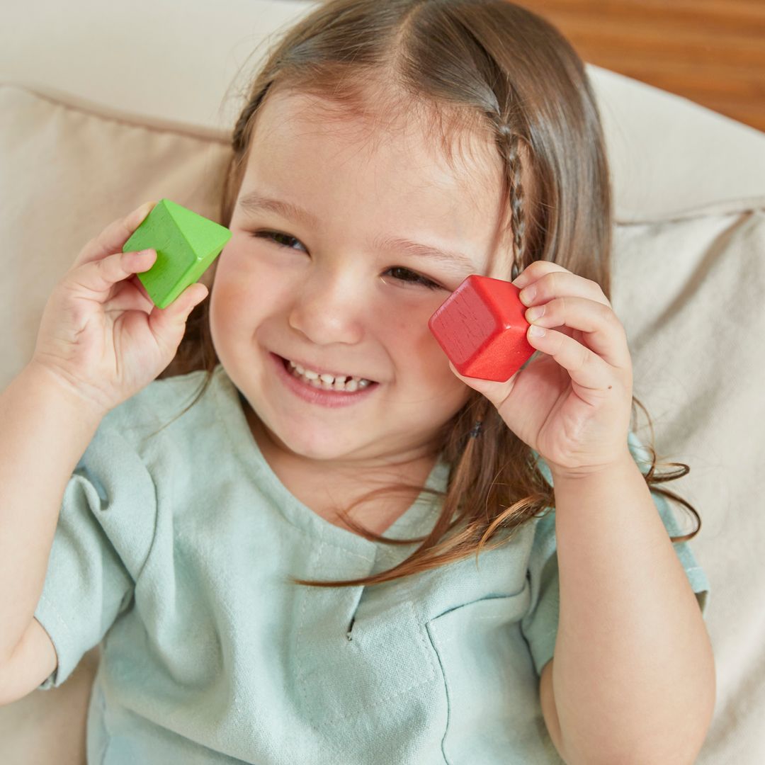 A happy toddler girl smiling while holding the green triangle and red square blocks from her PlanToys sorting set.