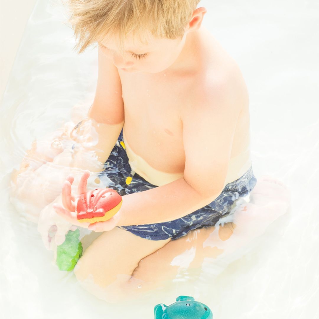 A toddler boy developing fine motor skills by watching water drain from the PlanToys red shell toy in the bathtub.