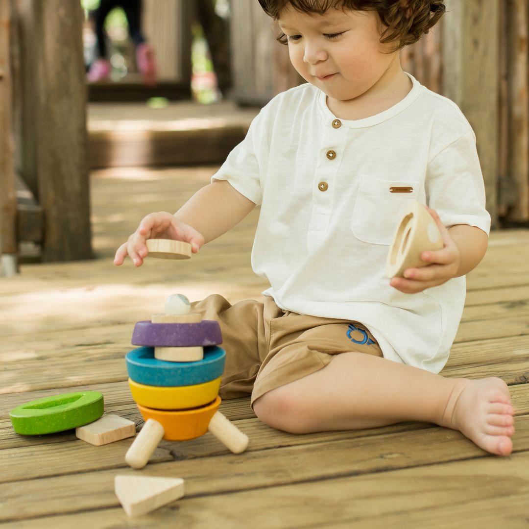 A young boy sitting on a wooden deck holding the nose cone of the colorful PlanToys stacking rocket toy.