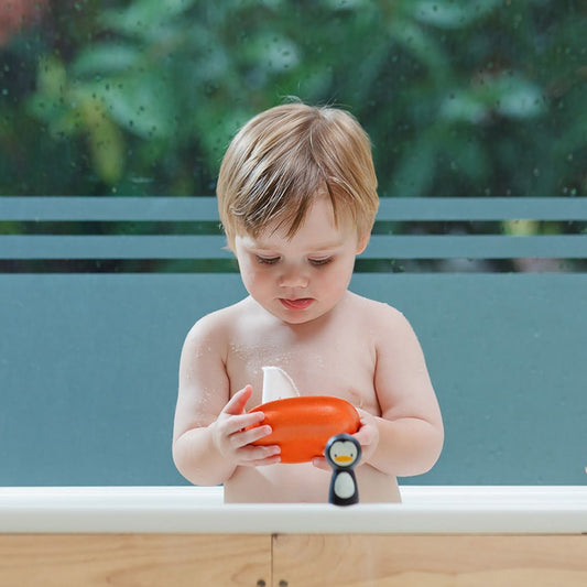 A toddler boy playing in the bathtub, holding the orange PlanToys boat while the wooden penguin rests on the edge.