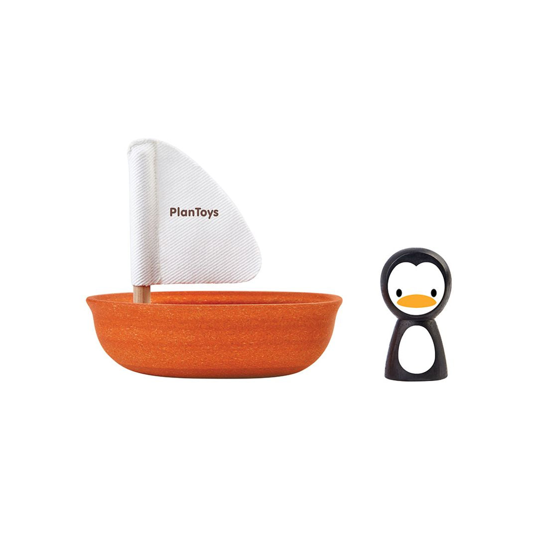 Isolated view of the orange PlanToys sailing boat with white sail next to the separate black and white penguin figure.