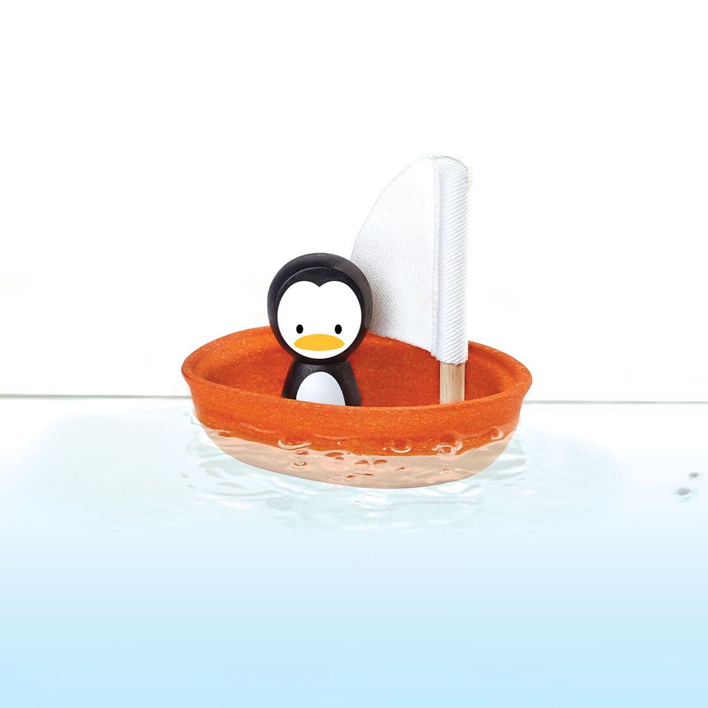 The sustainable PlanToys orange sailing boat floating in clear blue water with the penguin figure riding inside.