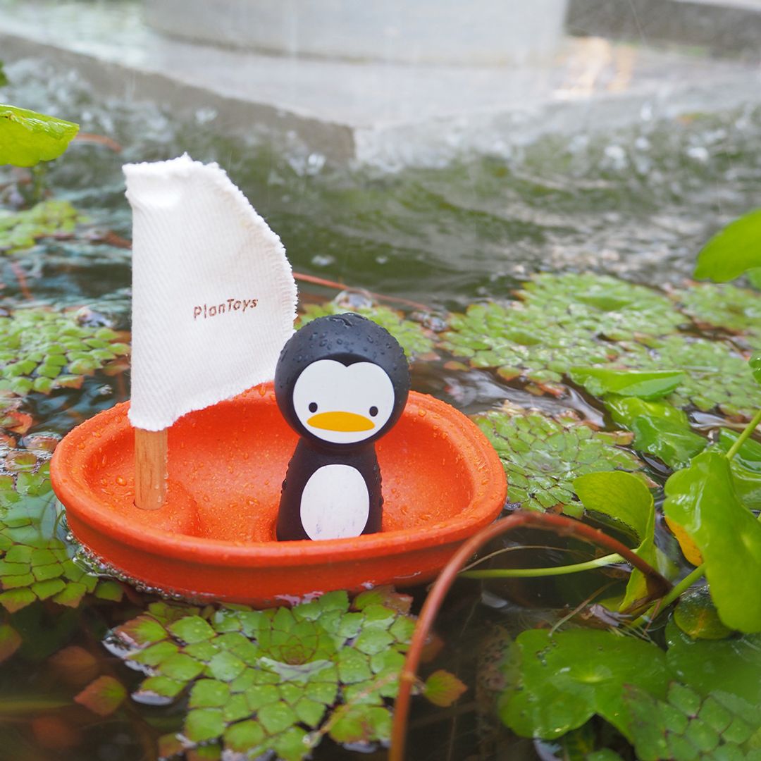 The earth-friendly PlanToys wooden sailing boat and penguin floating in an outdoor pond with green plants.