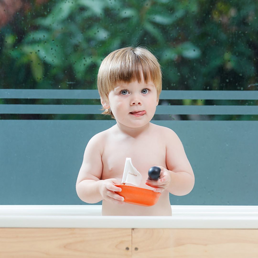 A young boy in the bathtub holding the orange PlanToys sailing boat in one hand and the penguin figure in the other.