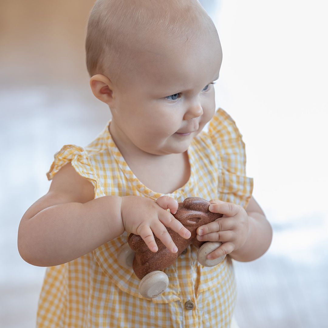 A happy baby in a yellow dress easily gripping the chemical-free brown PlanToys bunny toy in their hands.