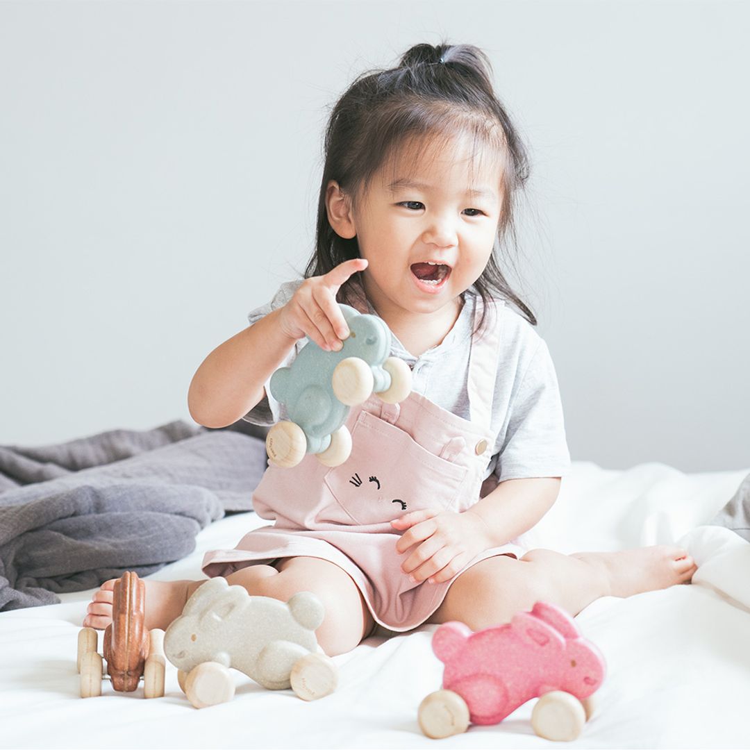 A happy toddler playing on the bed with their easy-to-grasp PlanToys grey wooden bunny toy.