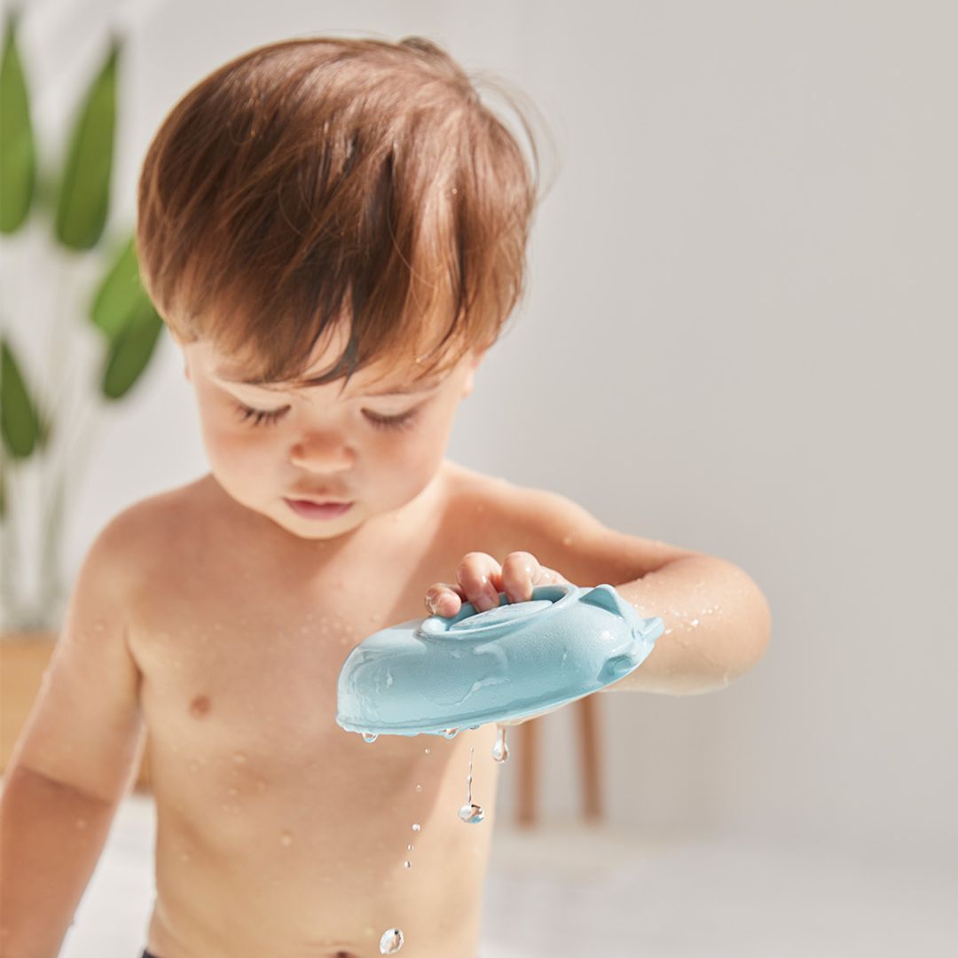 A toddler holding the tactile, pastel blue PlanToys rubber convertible boat during a sunny water play session.