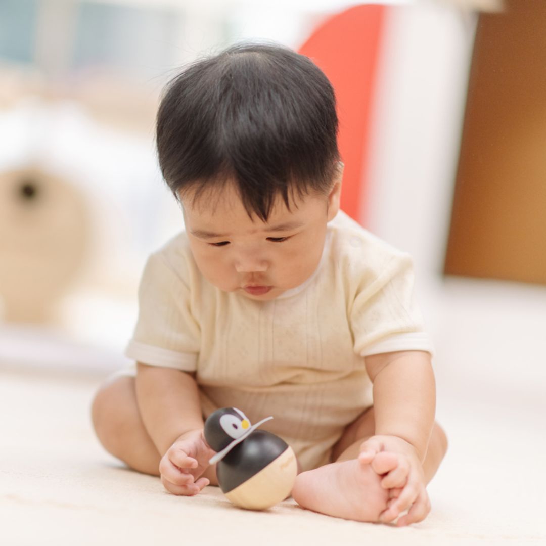 A baby sitting on a soft play mat looking down at the rocking PlanToys wooden penguin wobbler toy.