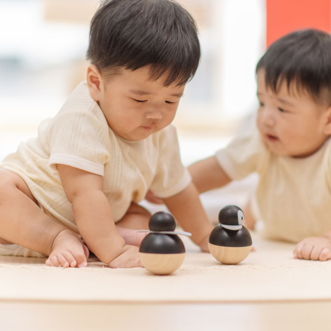 Two babies playing together on the floor with two eco-friendly PlanToys penguin wobbler toys.