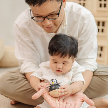 A father holding his baby in his lap while the baby grasps the durable PlanToys wooden penguin toy.