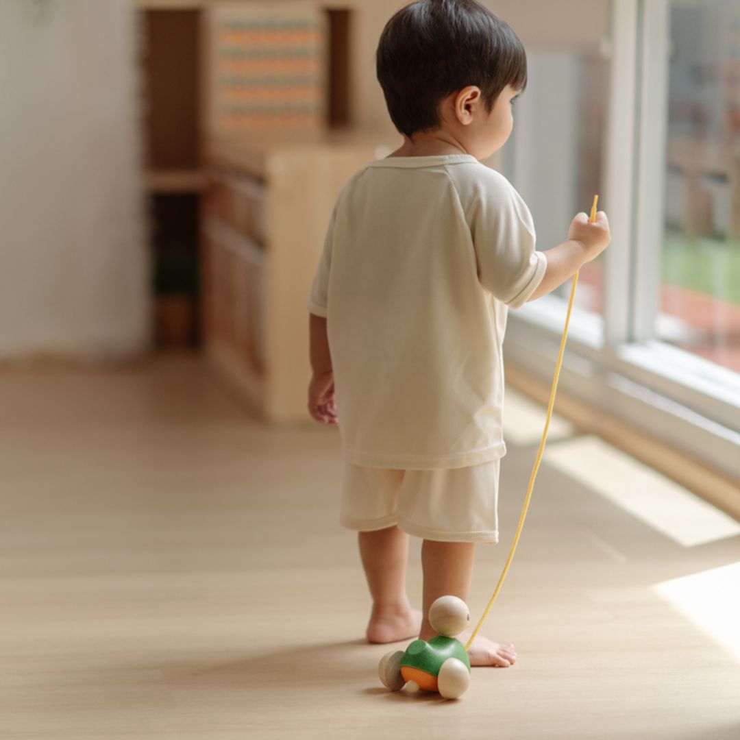 A little boy walking toward a bright window while pulling his classic wooden PlanToys duck companion.