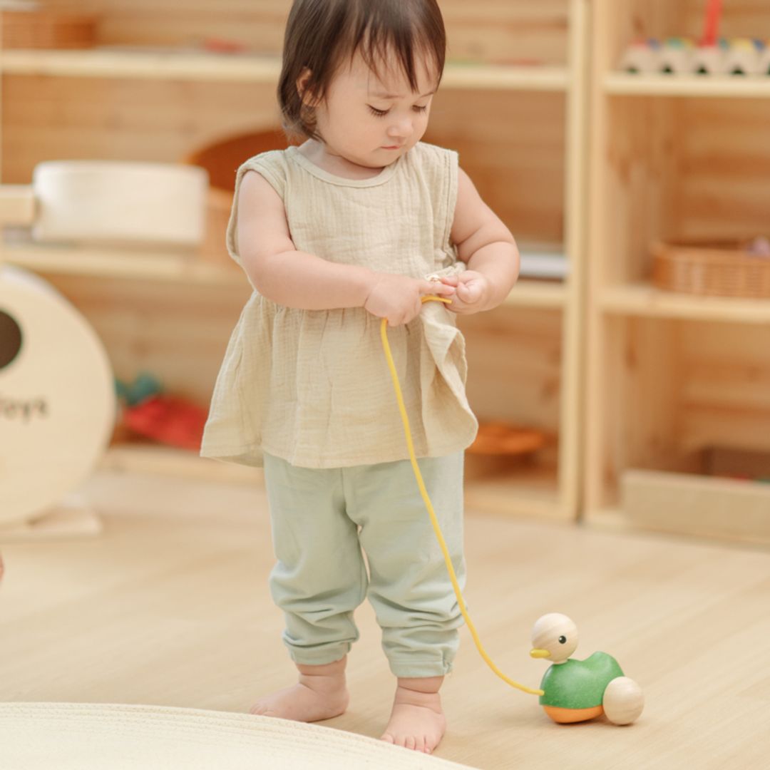 A toddler girl grasping the yellow string of the sustainable PlanToys wooden duck with both hands.
