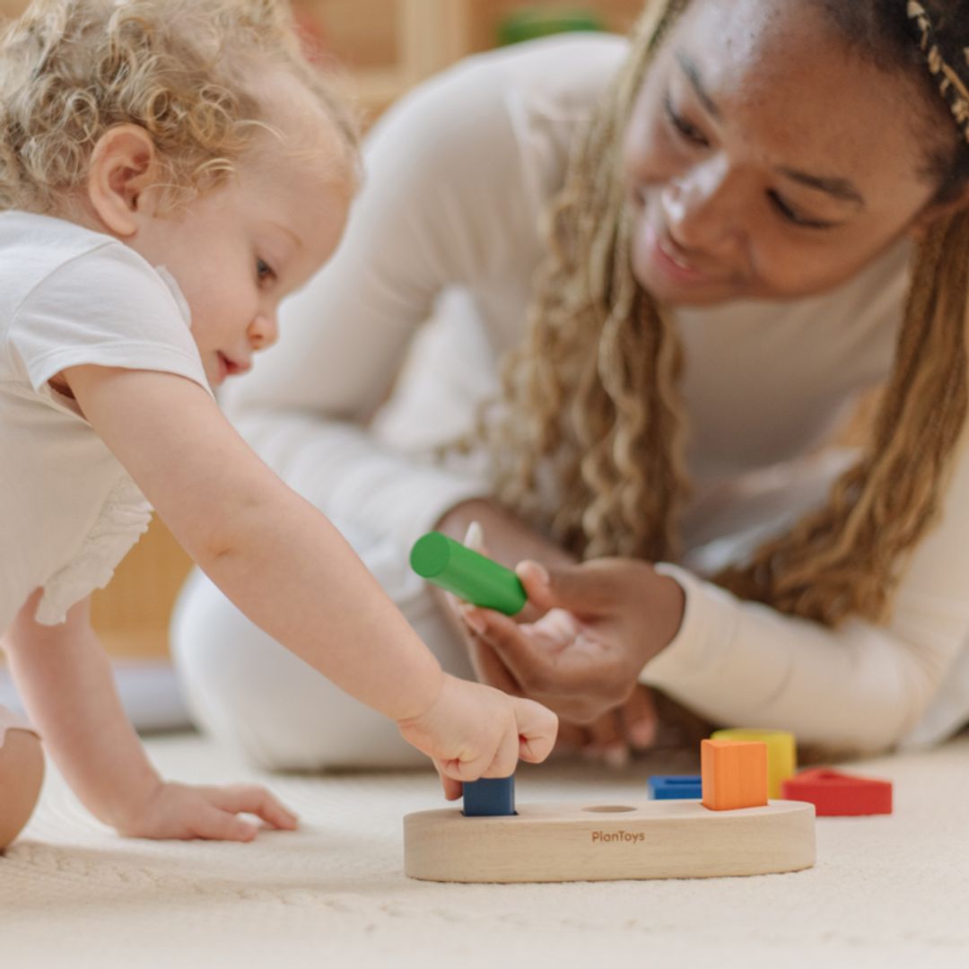 A crawling baby reaching for the sustainable PlanToys wooden sorting boat while an adult holds a green block.