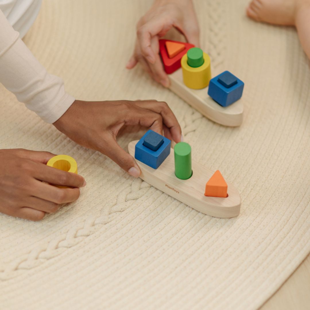 Overhead view of an adult guiding a child's hands to stack a blue wooden square onto the PlanToys boat peg.