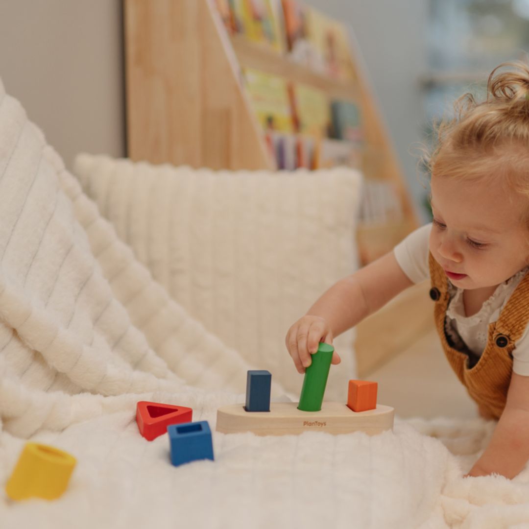 A young child practicing fine motor skills by holding a green cylinder piece from the PlanToys wooden shape sorter.