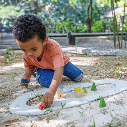 A little boy building a miniature city with his PlanToys flexible rubber road track in an outdoor sandbox.