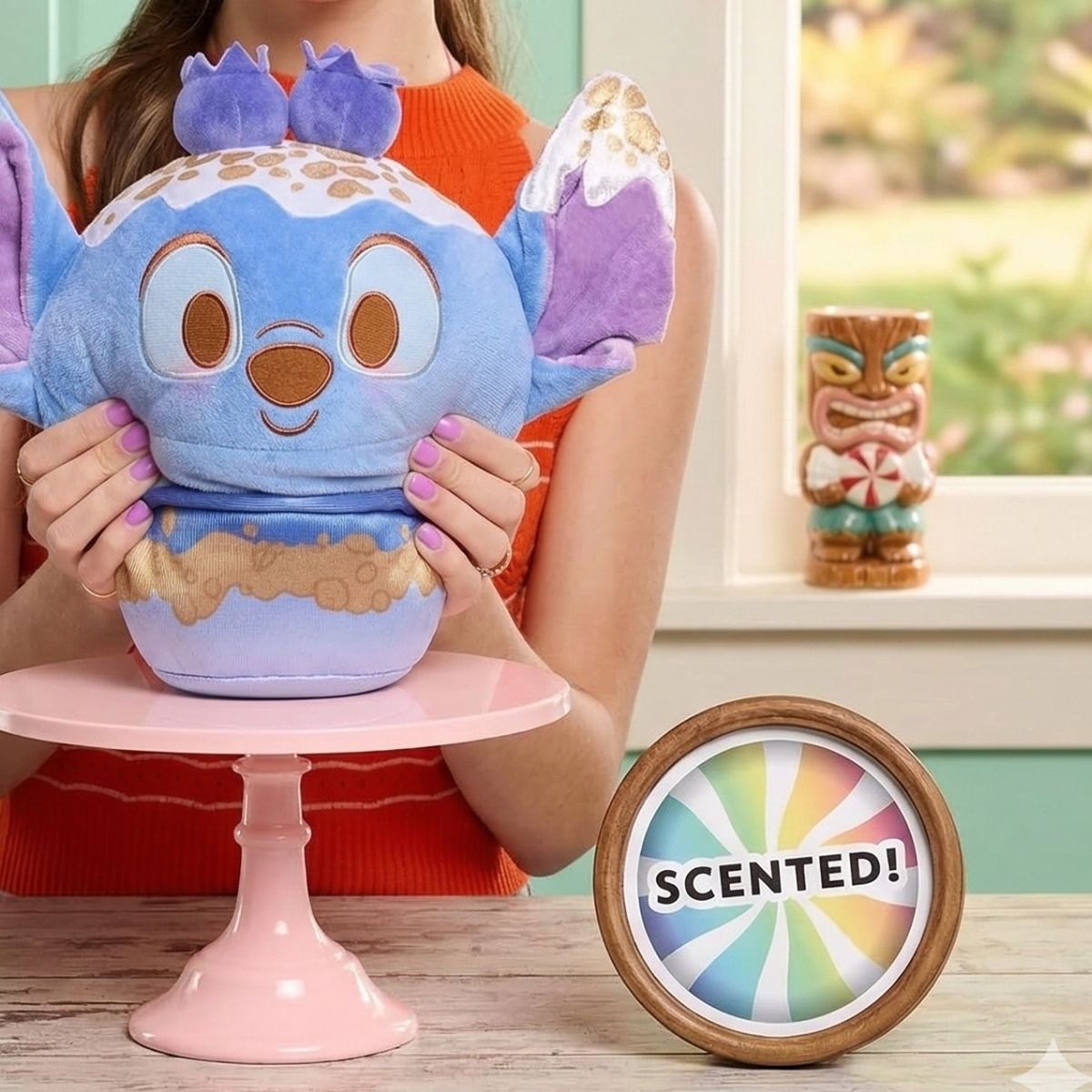 Lifestyle view of the scented Disney Munchlings Stitch plush resting on a pink cake stand, showcasing its fun food-inspired design.