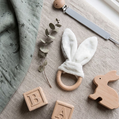 Happy Horse wooden bunny teether resting on a table next to wooden alphabet blocks, a green muslin blanket, and a pacifier clip.