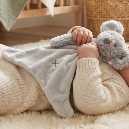 A baby cuddling the Happy Horse Mouse Mindy grey plush lovey closely while laying down on a soft cream rug.