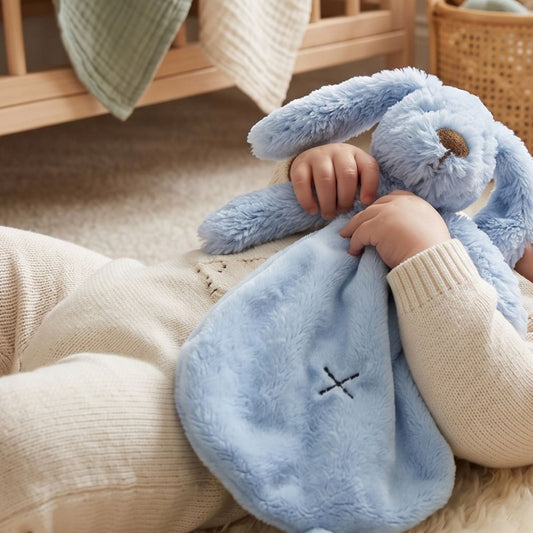 A baby cuddling the Happy Horse Rabbit Richie deep blue plush lovey closely while resting on a soft cream rug.