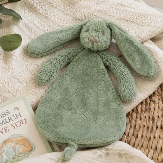 Happy Horse Rabbit Richie green plush lovey resting flat on a cozy knit blanket inside a nursery basket next to a baby book.