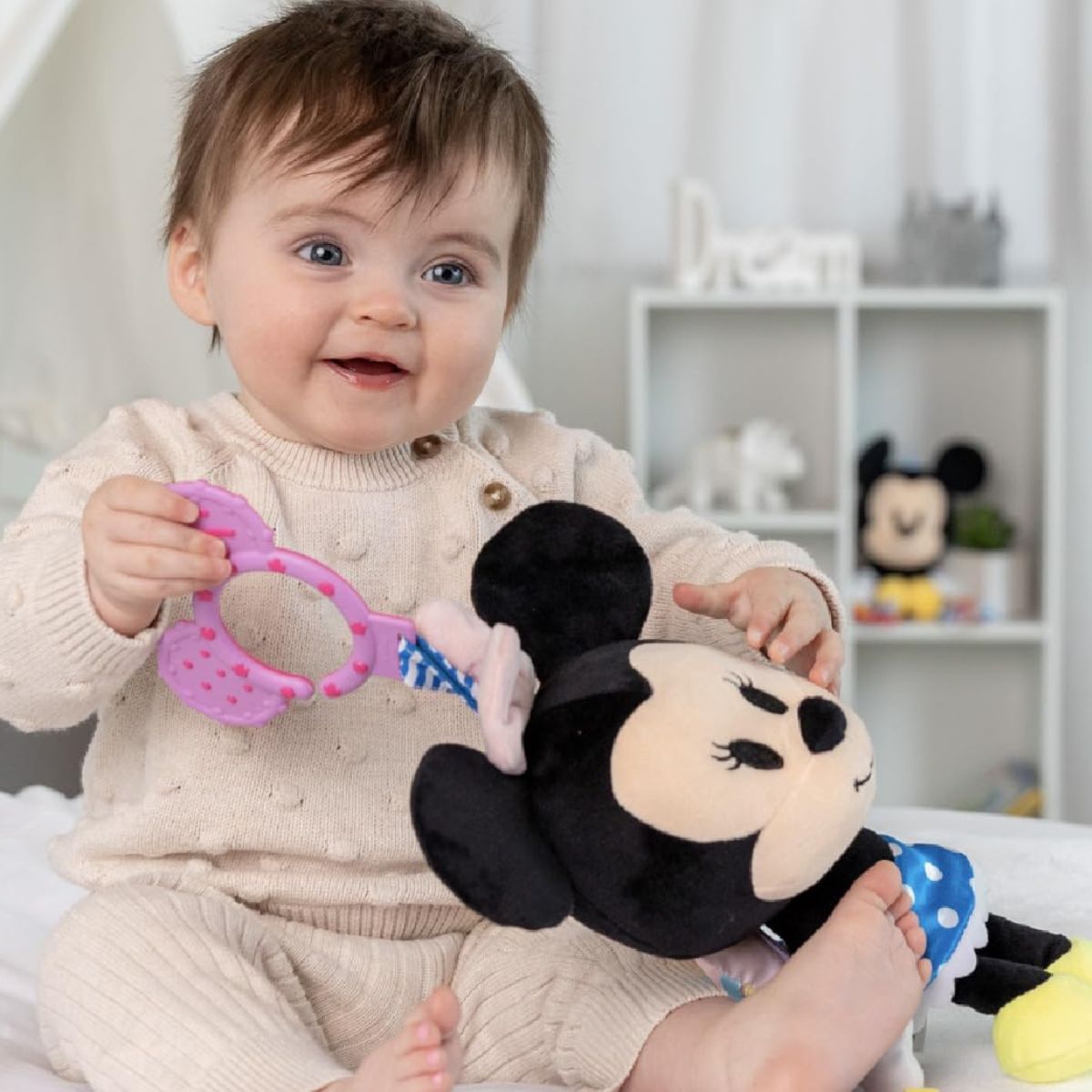 A happy baby sitting down on a plush floor mat while holding and exploring the large Minnie Mouse sensory activity plush.