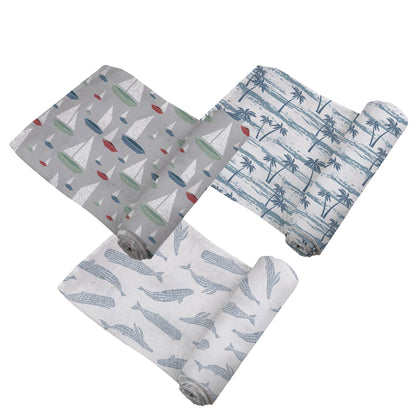 Top-down view of the three bamboo muslin swaddle blankets partially unrolled, featuring whale, sailboat, and palm tree prints.
