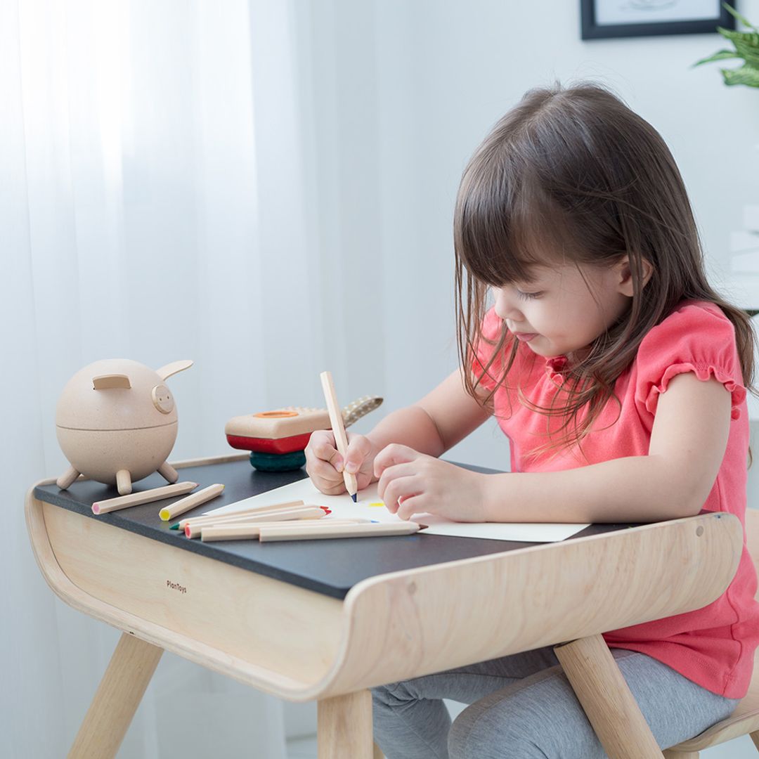 A little girl sitting comfortably in the ergonomic wooden chair and drawing on the versatile PlanToys kids table.