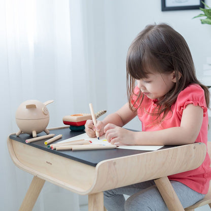 A little girl sitting comfortably in the ergonomic wooden chair and drawing on the versatile PlanToys kids table.