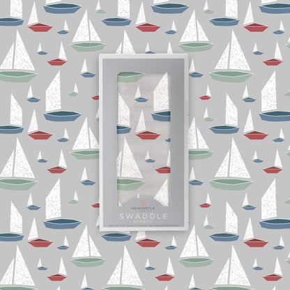 Flat lay of the Newcastle Classics Marina Sailboats bamboo muslin swaddle blanket shown with its branded grey packaging box in the center against a matching boat pattern background.