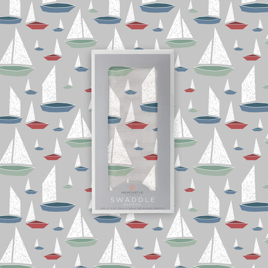 Flat lay of the Newcastle Classics Marina Sailboats bamboo muslin swaddle blanket shown with its branded grey packaging box in the center against a matching boat pattern background.