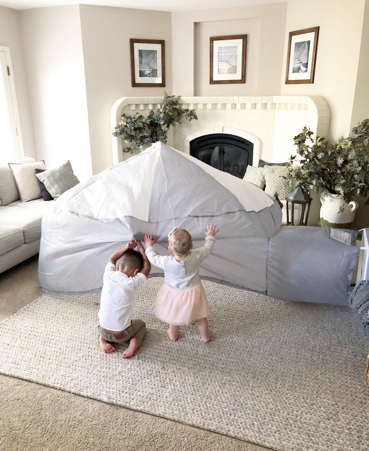 Two toddlers playfully exploring the outside of the inflated light gray and white AirFort in a cozy family room.