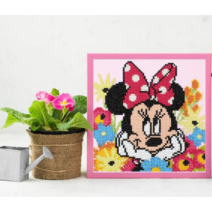 Completed Minnie Mouse diamond painting displayed in a pink frame sitting on a desk next to a potted pink plant and a mini silver watering can.